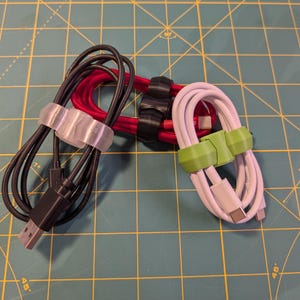 Puede incluir: Tres organizadores de cables de diferentes colores. Un organizador de cables negro con una correa de plástico transparente, un organizador de cables rojo con una correa de plástico negro y un organizador de cables blanco con una correa de plástico verde.