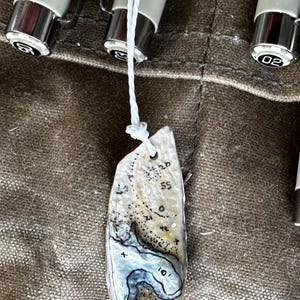 May include: An irregular, off-white shell pendant featuring a hand-drawn black ink topographic map design with contour lines and depth markers including 10, 20, 42, and 55. The pendant is suspended by a thin white string and positioned near three pens.