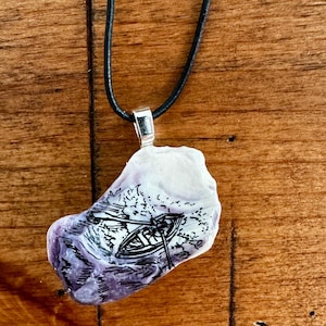 Może przedstawiać: Unikalny naszyjnik z wisiorkiem przedstawiający ręcznie malowaną scenę osoby wiosłującej w łodzi. Wisiorek ma nieregularny kształt z biało-fioletową kolorystyką, zawieszony na czarnym sznurku.