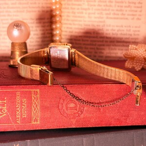May include: A gold-toned wristwatch with a rectangular case and a flexible metal expansion band. The watch features a thin safety chain attached to the clasp. The back of the watch case is metallic and rectangular, showing visible engraved text.