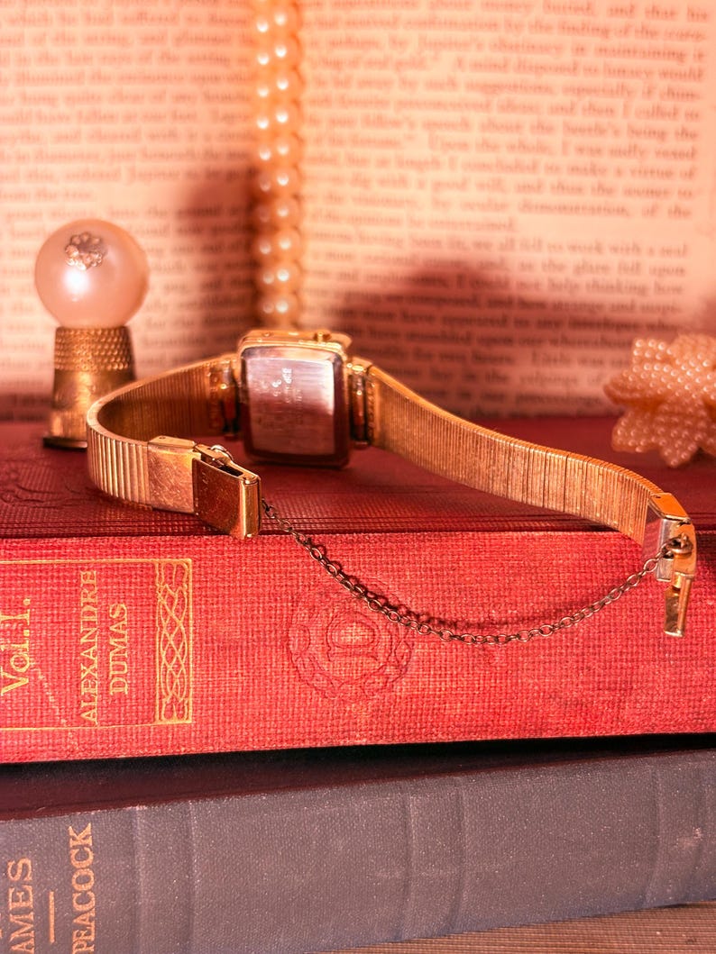 May include: A gold-toned wristwatch with a rectangular case and a flexible metal expansion band. The watch features a thin safety chain attached to the clasp. The back of the watch case is metallic and rectangular, showing visible engraved text.