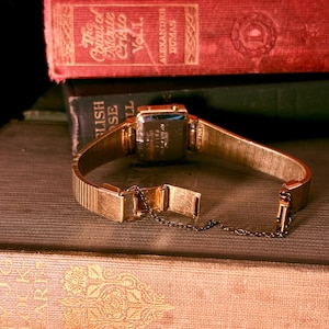 May include: A gold-tone wristwatch with a rectangular case and a flexible metal expansion band. The watch features a safety chain attached to the clasp. The back of the watch case displays engraved text including serial numbers and manufacturing details.