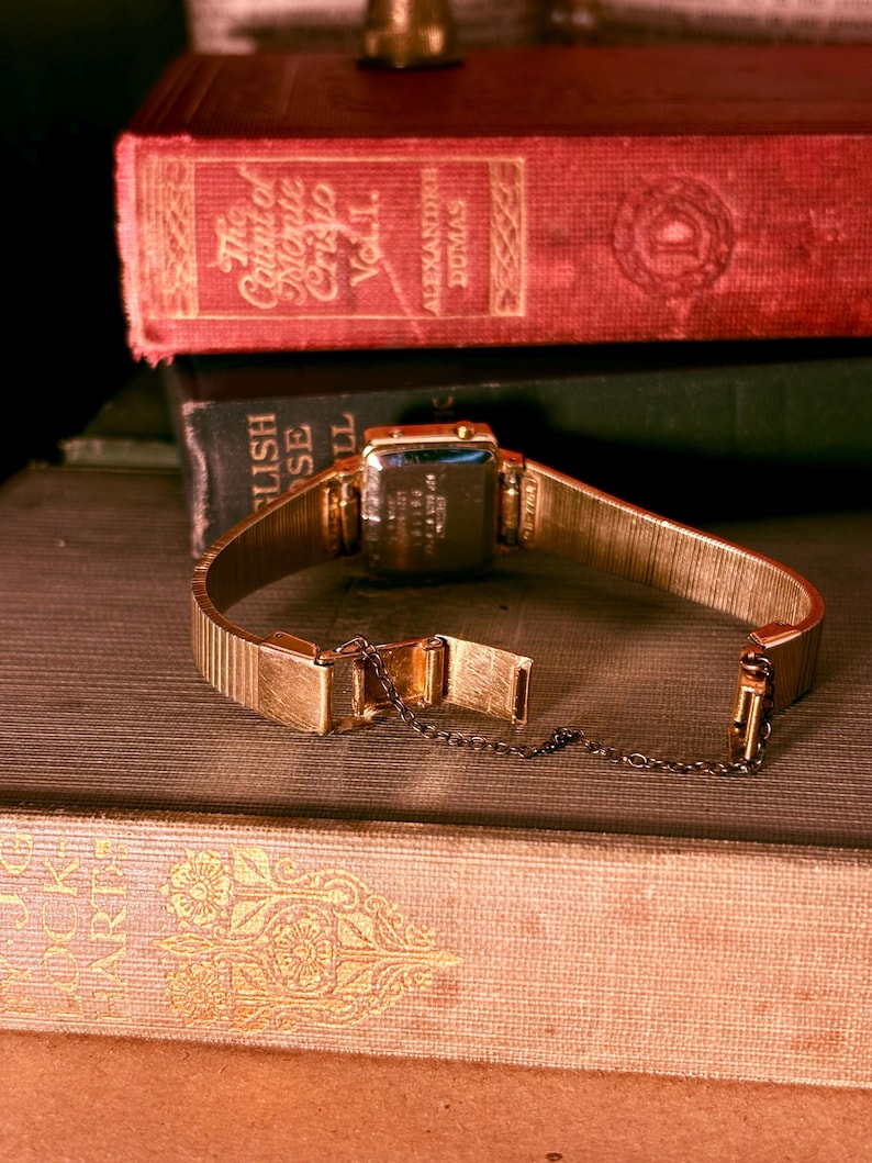 May include: A gold-tone wristwatch with a rectangular case and a flexible metal expansion band. The watch features a safety chain attached to the clasp. The back of the watch case displays engraved text including serial numbers and manufacturing details.