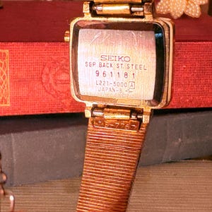 May include: A gold-tone Seiko wristwatch with a square case and a textured metal mesh strap. The back of the watch case is engraved with the text SEIKO, SGP BACK ST. STEEL, 961181, L221-5000 A, and JAPAN-B, showing a metallic finish and visible wear.
