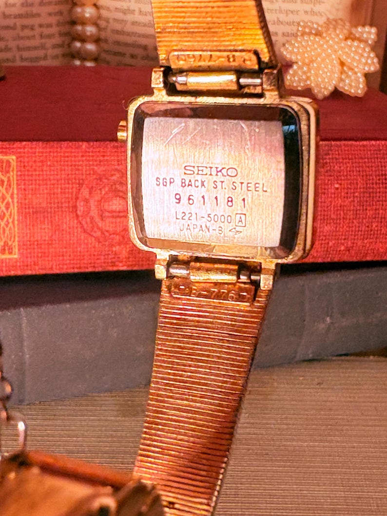 May include: A gold-tone Seiko wristwatch with a square case and a textured metal mesh strap. The back of the watch case is engraved with the text SEIKO, SGP BACK ST. STEEL, 961181, L221-5000 A, and JAPAN-B, showing a metallic finish and visible wear.