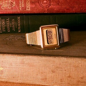 May include: A gold-tone digital wristwatch with a rectangular case and a textured metal link band. The watch face features a digital display with the text Seiko printed on the right side and a granular, metallic background texture behind the digits.