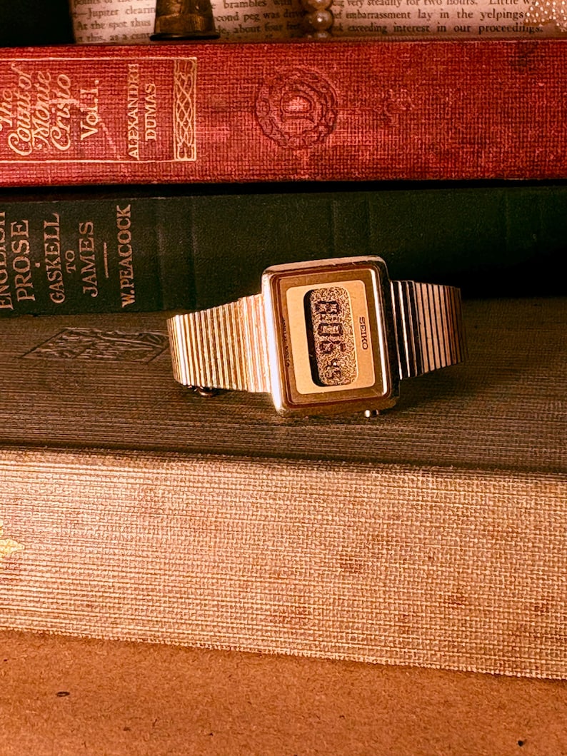 May include: A gold-tone digital wristwatch with a rectangular case and a textured metal link band. The watch face features a digital display with the text Seiko printed on the right side and a granular, metallic background texture behind the digits.