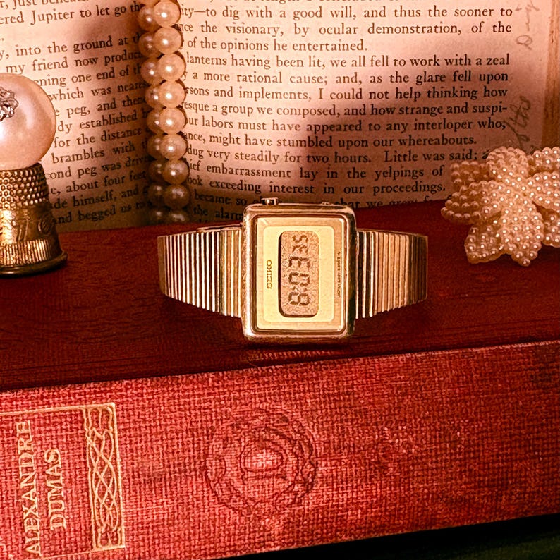 May include: A gold-tone digital wristwatch with a rectangular face and a textured, ribbed metal link band. The watch features a digital display showing the time, with the brand name Seiko printed vertically on the left side of the watch face.