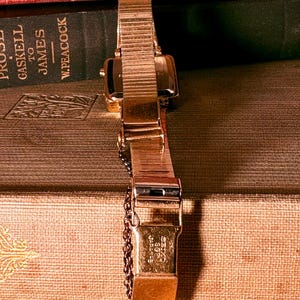 May include: A gold-tone wristwatch with a flexible metal expansion band and a safety chain. The clasp is engraved with the text SEIKO SGP JAPAN-G. The watch case is rectangular with a metallic finish, showing a classic link-style strap design.