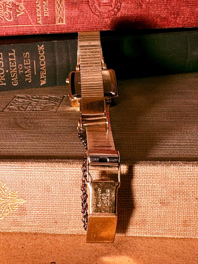 May include: A gold-tone wristwatch with a flexible metal expansion band and a safety chain. The clasp is engraved with the text SEIKO SGP JAPAN-G. The watch case is rectangular with a metallic finish, showing a classic link-style strap design.