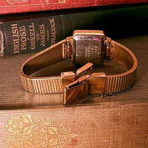 May include: A gold-tone wristwatch with a rectangular case and a flexible metal expansion band. The back of the watch case is engraved with the Seiko brand name, stainless steel markings, and serial numbers. The clasp is partially open.