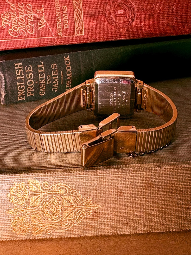 May include: A gold-tone wristwatch with a rectangular case and a flexible metal expansion band. The back of the watch case is engraved with the Seiko brand name, stainless steel markings, and serial numbers. The clasp is partially open.