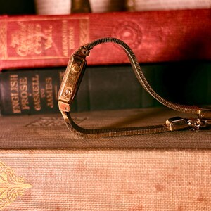 May include: A gold-toned wristwatch with a flexible, textured metal link band shaped into a heart. The watch features a rectangular case with visible side buttons and a metallic finish, resting against a stack of vintage hardcover books.