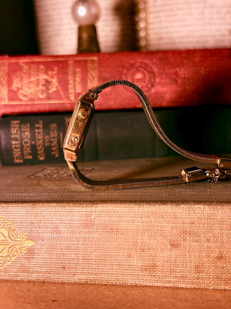 May include: A gold-toned wristwatch with a flexible, textured metal link band shaped into a heart. The watch features a rectangular case with visible side buttons and a metallic finish, resting against a stack of vintage hardcover books.
