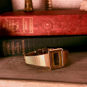 May include: A gold-tone digital wristwatch with a rectangular face and a textured, ribbed metal link band. The watch features a dark display screen with the Seiko brand name printed in small, clear lettering on the lower edge of the bezel.