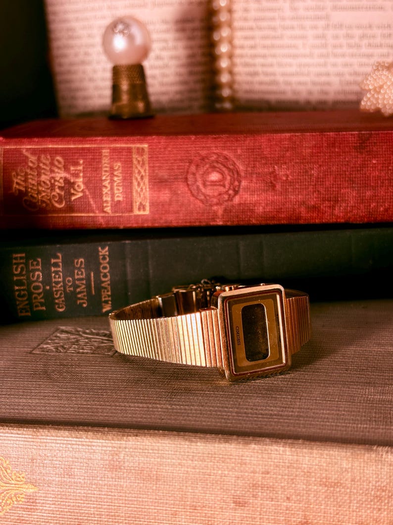 May include: A gold-tone digital wristwatch with a rectangular face and a textured, ribbed metal link band. The watch features a dark display screen with the Seiko brand name printed in small, clear lettering on the lower edge of the bezel.