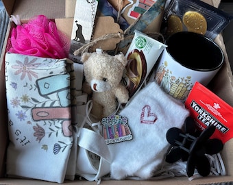May include: A gift box filled with various items, including a pink bath puff, a white tote bag with floral print and the text "It's a...", a small teddy bear, a white fuzzy sock with a red heart embroidered on it, a black hair clip, a mug with a book stack design, a chocolate bar, and a tea bag package.