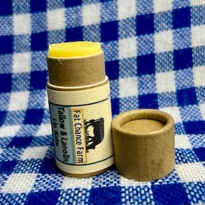 Puede incluir: Un tubo de bálsamo labial con bálsamo amarillo visible en la parte superior. La etiqueta dice "Fat Chance Farm Tallow & Lanolin Lip Balm" con un gráfico de vaca. Una tapa de tubo vacía separada está al lado.