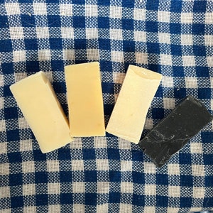 Puede incluir: Cuatro barras de jabón sobre una tela a cuadros azul y blanca. Dos barras son de color amarillo claro, una es de color amarillo más oscuro y una es negra.