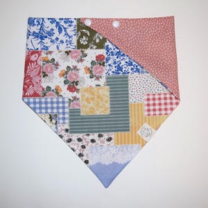 May include: A patchwork bandana with a variety of floral, geometric, and plaid patterns in shades of blue, pink, red, green, and yellow. The bandana is tied with a knot at the top and has two white buttons.