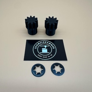 Könnte beinhalten: Zwei schwarze zahnradförmige Kunststoffbauteile, zwei runde Sternscheiben aus Metall und eine rechteckige schwarze Karte mit dem weißen Text SCHMELZKAMMER IHRE IDEE IN 3D und einem Symbol einer 3D-Druckerdüse.