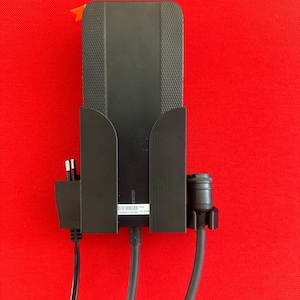 May include: Black electronic device with a textured surface, mounted on a red background. The device has a power adapter and a cable with a connector. An orange tab is visible at the top.