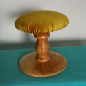 Puede incluir: Un pequeño taburete decorativo con un cojín redondo de terciopelo amarillo mostaza. El cojín se asienta sobre un pedestal de madera torneada, que está unido a una base redonda de madera. El taburete está sobre una superficie turquesa y una pared blanca.