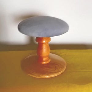 May include: A wooden hat stand with a round, light blue padded top. The stand has a turned wooden stem and a circular base. The wood has a warm, natural finish, and the stand is set against a yellow background.