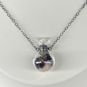 May include: A clear glass pendant necklace with a silver chain. The pendant is a small, round bottle filled with purple and pink stones and small green stems. The bottle has a silver cap and a clear, faceted stopper.