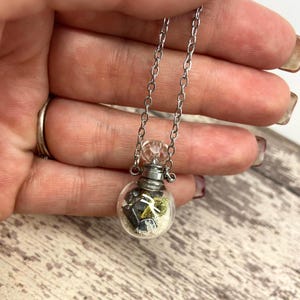 Op de afbeelding: Een zilveren ketting met een kleine, heldere glazen flaconhanger. De flacon bevat kleine, donkere stenen, zand en groene elementen. De ketting heeft een fijne zilveren ketting met een heldere kristallen kraal aan de bovenkant van de flacon.