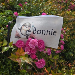 May include: A rectangular off-white canvas zipped pouch featuring a printed illustration of a small grey orca whale surrounded by blue bubbles and red coral, with the name Bonnie written in black script lettering on the right side.