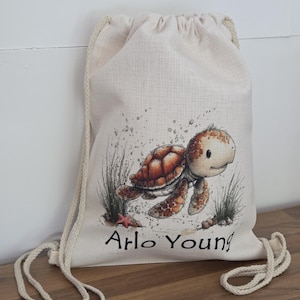 May include: A beige drawstring bag with a cartoon turtle illustration in brown and orange, and the text "Arlo Young". The bag has a cream-colored drawstring closure and is made of a textured fabric.