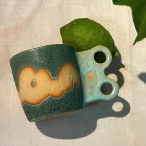 Puede incluir: Una taza de cerámica turquesa con un diseño de asa único y un patrón floral beige. Una lima verde descansa sobre el borde. La taza está sobre una superficie blanca.