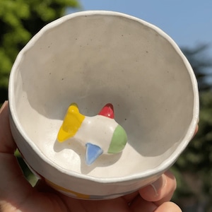 Puede incluir: Un tazón de cerámica blanco con una figurita de avión colorida en su interior. El avión está pintado de amarillo, rojo, verde y azul.