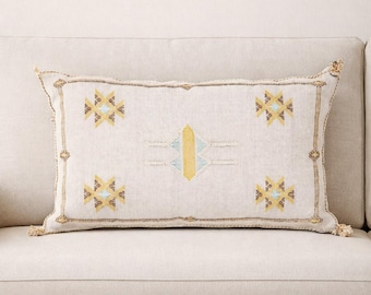 May include: A rectangular throw pillow in a light beige fabric featuring embroidered geometric patterns in yellow, light blue, and brown. The design includes four star-like motifs at the corners and a central vertical yellow bar.