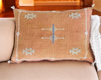 May include: A rectangular throw pillow with a light brown background and a geometric pattern in shades of blue, white, and beige. The pillow has a fringe trim.