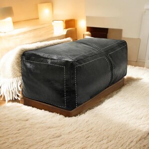 May include: Black leather ottoman with white stitching and a wooden base. The ottoman is sitting on a white shag rug.