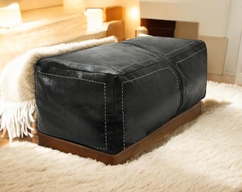 May include: Black leather ottoman with white stitching and a wooden base. The ottoman is sitting on a white shag rug.