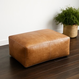 May include: A rectangular, light brown leather ottoman. The ottoman has a stitched design and is placed on a dark wood floor. A woven basket with a green plant is in the background.