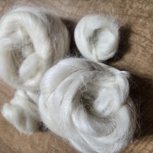 May include: Close-up of white fluffy fiber, likely wool or cotton, arranged in three loose balls. The fiber is soft and appears to be of high quality.