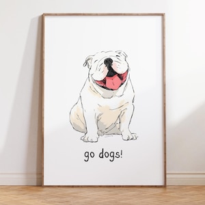 May include: A framed print showing a smiling white bulldog illustration with a pink tongue. The artwork has black outlines and the words "go dogs!" in black script at the bottom. The frame is a light brown colour.