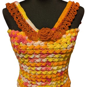 Peut inclure: Un haut crocheté avec un motif coloré et multicolore dans des tons de rose, jaune, orange et blanc. Le haut est doté d'une bordure en dentelle crochetée autour de l'encolure et d'une fleur en crochet en guise d'embellissement au centre.