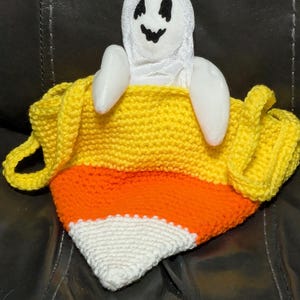 May include: A handmade Halloween decoration featuring a plush ghost emerging from a knitted candy corn-shaped container. The ghost is white with a black smiling face. The candy corn is yellow, orange, and white.
