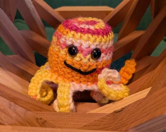 Può includere: Un giocattolo a forma di polpo all'uncinetto nei toni dell'arancione, giallo e rosa, con un volto sorridente. Il polpo è posto in una ciotola decorativa di legno. Lo sfondo è in tessuto verde.