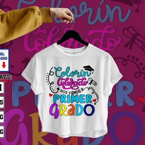 Puede incluir: Camiseta blanca con el texto "Colorín Colorado Nos Vamos a Primer Grado" en letras coloridas. El diseño incluye un gorro de graduación, un pergamino y corazones. La camiseta está colgada en una percha negra.