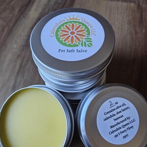 May include: A silver tin with a label that reads "Calendula Queen LLC" and "Pet Safe Salve". The tin contains a yellow salve. The label also lists the ingredients: olive oil, calendula, shea butter, and beeswax. The label also states that the salve is manufactured by Calendula Queen LLC and includes the address 595 S Perry Pkwy 53575.