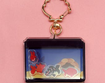 May include: A clear acrylic keychain featuring a miniature aquarium scene with colorful goldfish. The keychain has a gold-colored clasp and a rectangular design. The aquarium scene includes a red, black and white, and pink goldfish.