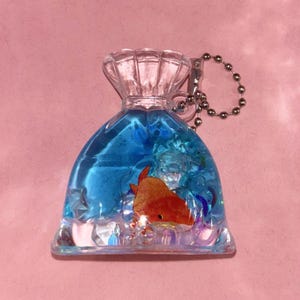 May include: A clear, fish-shaped keychain filled with blue liquid and a small orange fish figurine. The keychain has a silver chain and a clear, textured top. The background is a solid pink color.