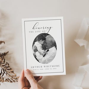 Puede incluir: Una tarjeta conmemorativa en blanco y negro con el texto "honoring the life of" sobre una foto ovalada de un hombre y un ni&ntilde;o. El nombre "Arthur Whitmore" y las fechas est&aacute;n debajo de la foto. La tarjeta est&aacute; sostenida por una mano.