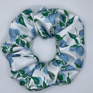 Op de afbeelding: Witte satijnen scrunchie met een blauw hortensia bloemenpatroon. De scrunchie heeft een herhalend ontwerp van blauwe hortensiabloemen en groene bladeren. De stof heeft een lichte glans, wat bijdraagt aan de elegante uitstraling. Dit haaraccessoire is geschikt voor diverse kapsels.